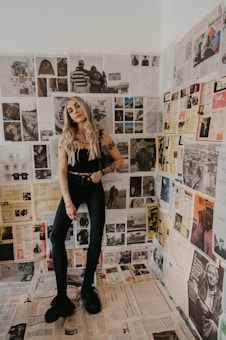 A woman with long blonde hair and tattoos stands in a room covered with newspaper clippings on the walls and floor. She is wearing a black sleeveless top, dark jeans, and black shoes. The clippings feature various images and text in different styles, showcasing a vintage and eclectic aesthetic.