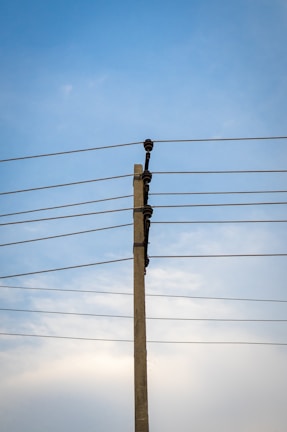 A concrete utility pole supports several electrical wires extending horizontally against a clear blue sky. The wires are evenly spaced and attached using insulators on the pole.