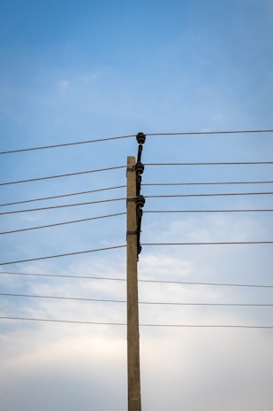 A concrete utility pole supports several electrical wires extending horizontally against a clear blue sky. The wires are evenly spaced and attached using insulators on the pole.