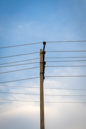 A concrete utility pole supports several electrical wires extending horizontally against a clear blue sky. The wires are evenly spaced and attached using insulators on the pole.