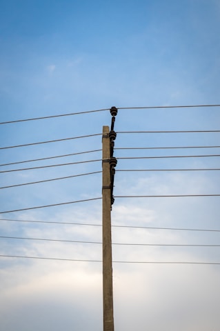 A concrete utility pole supports several electrical wires extending horizontally against a clear blue sky. The wires are evenly spaced and attached using insulators on the pole.