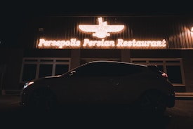 A neon-lit sign for Persepolis Persian Restaurant is prominently displayed above the entrance. The outline of a car is visible in shadow in front of the building, contributing to a nighttime ambiance.