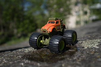 A toy monster truck with large, rugged wheels and an orange and green color scheme is positioned on a rock surface. The background features a blurred natural setting with greenery.