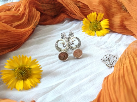 Intricately designed earrings set against a white fabric backdrop, surrounded by bright yellow flowers and draped with a flowing orange cloth.