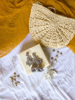 A textured crochet handbag is resting on a mustard yellow fabric. Below the bag is a white cloth with a floral embroidery design. On top of the white cloth, there are silver ornate earrings placed on a small square box. There are also small shells scattered around the cloth.