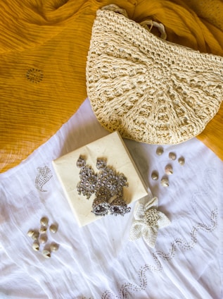 A textured crochet handbag is resting on a mustard yellow fabric. Below the bag is a white cloth with a floral embroidery design. On top of the white cloth, there are silver ornate earrings placed on a small square box. There are also small shells scattered around the cloth.
