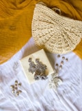 A textured crochet handbag is resting on a mustard yellow fabric. Below the bag is a white cloth with a floral embroidery design. On top of the white cloth, there are silver ornate earrings placed on a small square box. There are also small shells scattered around the cloth.