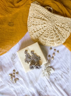 A textured crochet handbag is resting on a mustard yellow fabric. Below the bag is a white cloth with a floral embroidery design. On top of the white cloth, there are silver ornate earrings placed on a small square box. There are also small shells scattered around the cloth.