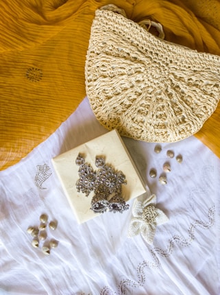 A textured crochet handbag is resting on a mustard yellow fabric. Below the bag is a white cloth with a floral embroidery design. On top of the white cloth, there are silver ornate earrings placed on a small square box. There are also small shells scattered around the cloth.