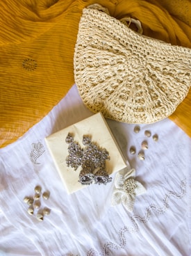 A textured crochet handbag is resting on a mustard yellow fabric. Below the bag is a white cloth with a floral embroidery design. On top of the white cloth, there are silver ornate earrings placed on a small square box. There are also small shells scattered around the cloth.