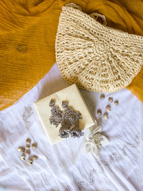 A textured crochet handbag is resting on a mustard yellow fabric. Below the bag is a white cloth with a floral embroidery design. On top of the white cloth, there are silver ornate earrings placed on a small square box. There are also small shells scattered around the cloth.