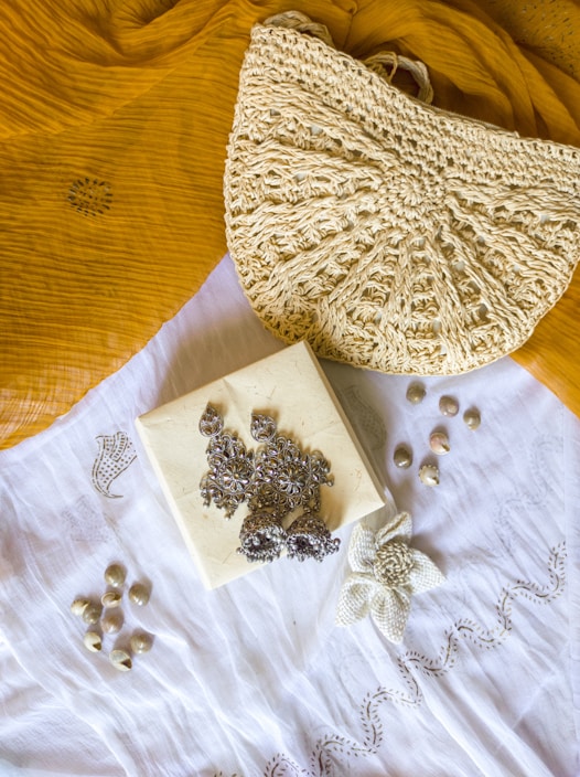A textured crochet handbag is resting on a mustard yellow fabric. Below the bag is a white cloth with a floral embroidery design. On top of the white cloth, there are silver ornate earrings placed on a small square box. There are also small shells scattered around the cloth.