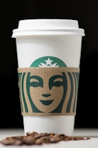 A white coffee cup with a cardboard sleeve featuring a green logo design, accompanied by scattered coffee beans in the foreground against a dark background.