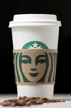 A white coffee cup with a cardboard sleeve featuring a green logo design, accompanied by scattered coffee beans in the foreground against a dark background.