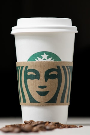 A white coffee cup with a cardboard sleeve featuring a green logo design, accompanied by scattered coffee beans in the foreground against a dark background.