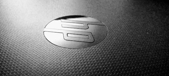 A close-up shot of a textured surface featuring a shiny, circular logo with stylized letters in the center. The logo appears to contrast with the matte surface, creating a sleek and modern aesthetic.