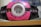 A pink and black electronic device with various symbols and a circular transparent screen in the center. There is a green light above the screen and multiple circular pink buttons on either side of the device. The device appears to have a barcode visible through the transparent screen.