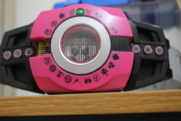 A pink and black electronic device with various symbols and a circular transparent screen in the center. There is a green light above the screen and multiple circular pink buttons on either side of the device. The device appears to have a barcode visible through the transparent screen.
