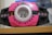 A pink and black electronic device with various symbols and a circular transparent screen in the center. There is a green light above the screen and multiple circular pink buttons on either side of the device. The device appears to have a barcode visible through the transparent screen.
