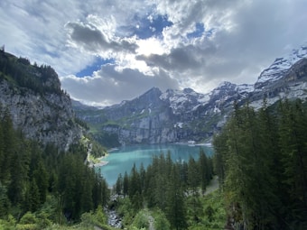 A serene alpine landscape featuring a large turquoise lake surrounded by dense forests and towering snow-capped mountains. The sky is partly cloudy, with sunlight breaking through, creating dramatic lighting effects.