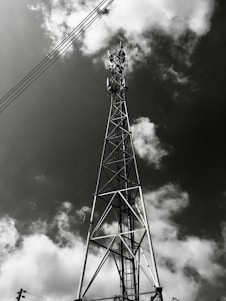 A sleek black and white photo of a cell tower against a cloudy sky, highlighting its structural details.