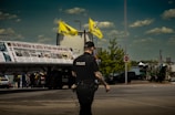 A police officer with visible tattoos is standing on the street, facing away from the camera. Two yellow flags with 'Khalistan' written on them are prominently displayed above a large banner on a truck. The sky is clear with some clouds and there are vehicles and people in the background.