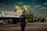 A police officer with visible tattoos is standing on the street, facing away from the camera. Two yellow flags with 'Khalistan' written on them are prominently displayed above a large banner on a truck. The sky is clear with some clouds and there are vehicles and people in the background.