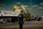 A police officer with visible tattoos is standing on the street, facing away from the camera. Two yellow flags with 'Khalistan' written on them are prominently displayed above a large banner on a truck. The sky is clear with some clouds and there are vehicles and people in the background.