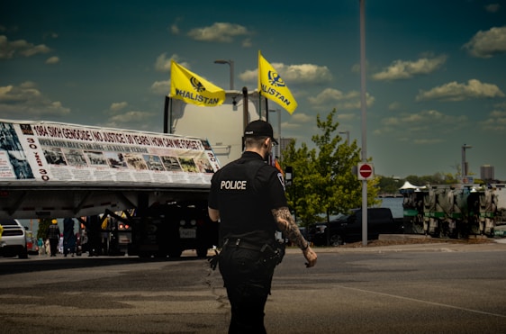 A police officer with visible tattoos is standing on the street, facing away from the camera. Two yellow flags with 'Khalistan' written on them are prominently displayed above a large banner on a truck. The sky is clear with some clouds and there are vehicles and people in the background.