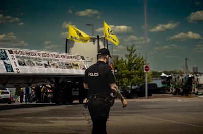A police officer with visible tattoos is standing on the street, facing away from the camera. Two yellow flags with 'Khalistan' written on them are prominently displayed above a large banner on a truck. The sky is clear with some clouds and there are vehicles and people in the background.