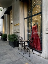 A stylish storefront displays a vibrant red dress on a mannequin behind a glass window with ornate metal latticework. Two intricately designed metal chairs and a small table are placed on the sidewalk, next to potted plants lining the exterior. The establishment name is visible in gold letters on the stone facade.