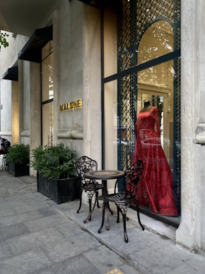A stylish storefront displays a vibrant red dress on a mannequin behind a glass window with ornate metal latticework. Two intricately designed metal chairs and a small table are placed on the sidewalk, next to potted plants lining the exterior. The establishment name is visible in gold letters on the stone facade.