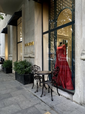 A stylish storefront displays a vibrant red dress on a mannequin behind a glass window with ornate metal latticework. Two intricately designed metal chairs and a small table are placed on the sidewalk, next to potted plants lining the exterior. The establishment name is visible in gold letters on the stone facade.