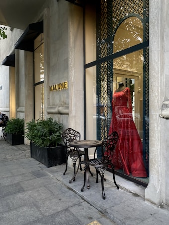 A stylish storefront displays a vibrant red dress on a mannequin behind a glass window with ornate metal latticework. Two intricately designed metal chairs and a small table are placed on the sidewalk, next to potted plants lining the exterior. The establishment name is visible in gold letters on the stone facade.