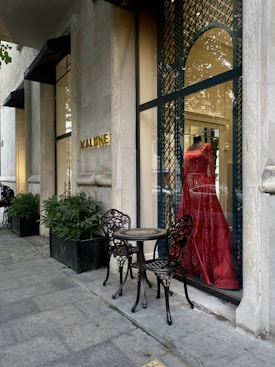 A stylish storefront displays a vibrant red dress on a mannequin behind a glass window with ornate metal latticework. Two intricately designed metal chairs and a small table are placed on the sidewalk, next to potted plants lining the exterior. The establishment name is visible in gold letters on the stone facade.