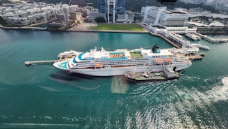 A large cruise ship docked at a port with clear turquoise water surrounding it. The ship has multiple decks, swimming pools, and recreational areas visible on the top. Adjacent to the ship, a smaller vessel is moored. In the background, there's a modern urban area with high-rise buildings, office complexes, and green spaces.