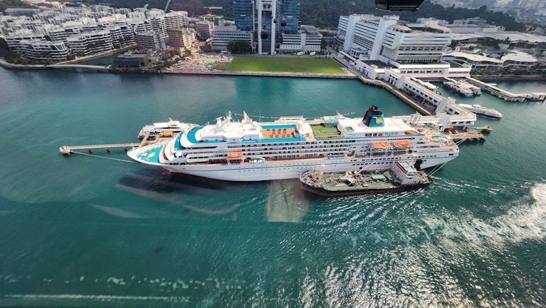 A large cruise ship docked at a port with clear turquoise water surrounding it. The ship has multiple decks, swimming pools, and recreational areas visible on the top. Adjacent to the ship, a smaller vessel is moored. In the background, there's a modern urban area with high-rise buildings, office complexes, and green spaces.