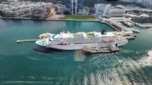 A large cruise ship docked at a port with clear turquoise water surrounding it. The ship has multiple decks, swimming pools, and recreational areas visible on the top. Adjacent to the ship, a smaller vessel is moored. In the background, there's a modern urban area with high-rise buildings, office complexes, and green spaces.