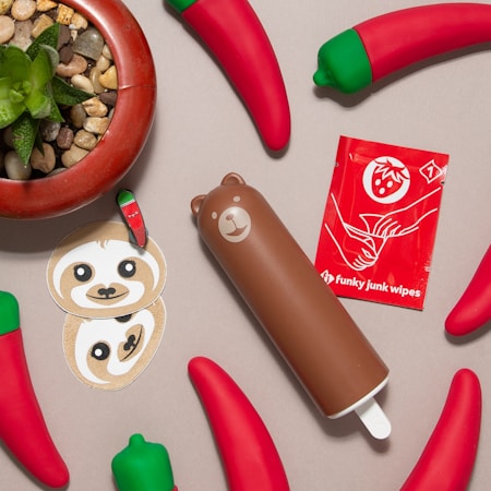 A collection of brightly colored objects is arranged on a beige surface. There is a brown bear-shaped container, multiple red chili pepper-shaped items with green caps, a small pot with a succulent plant and pebbles, two cartoonish sloth face stickers, a mini chili pepper sticker, and a red packet labeled 'Funky junk wipes' with an illustration of hands and a strawberry.
