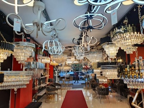 A well-lit interior showroom with a variety of modern and ornate chandeliers and light fixtures. The walls are red and black, and the floor is shiny white tile. There are chairs, small tables, and various decor items throughout the space. A red carpet runs down the center towards a window displaying outdoor scenery.