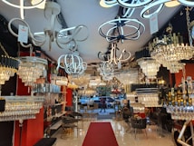 A well-lit interior showroom with a variety of modern and ornate chandeliers and light fixtures. The walls are red and black, and the floor is shiny white tile. There are chairs, small tables, and various decor items throughout the space. A red carpet runs down the center towards a window displaying outdoor scenery.