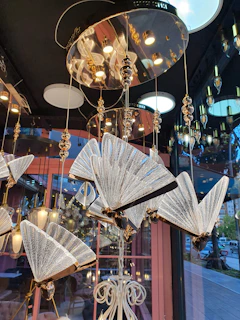 An elaborate chandelier with multiple fan-shaped light fixtures made of glass and metal. The fixtures hang from a glossy, reflective surface with gold accents, creating an intricate and luxurious design. The background shows a glimpse of a window reflecting outdoor elements.