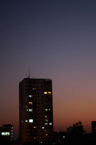 A sleek, modern luxury apartment building at dusk with warm golden lights glowing against a navy blue sky.