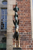 A statue of stacked animals stands against a red brick wall. The sculpture features a donkey at the bottom, a dog on its back, a cat on the dog's back, and a rooster perched on top.