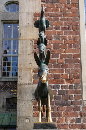 A statue of stacked animals stands against a red brick wall. The sculpture features a donkey at the bottom, a dog on its back, a cat on the dog's back, and a rooster perched on top.