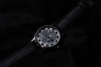A detailed mechanical watch is displayed on a textured black surface. The watch features visible gears and a complex design on the face, surrounded by a metal rim. The strap is made of black leather with an embossed pattern, complementing the sophisticated appearance of the watch.
