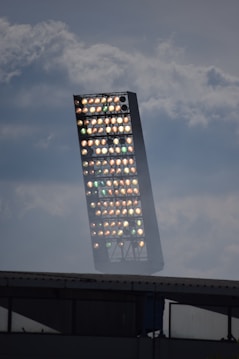 A large outdoor floodlight structure with numerous individual lights against a backdrop of cloudy sky. The floodlight tower appears slightly angled and is set above the roof of a building.