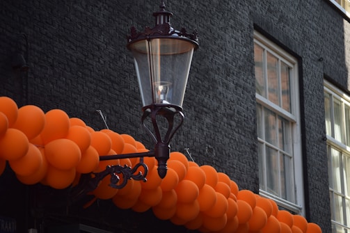 A cluster of vibrant orange balloons is arranged in front of an old-fashioned street lamp mounted on a black, textured brick wall. Two windows with white frames are visible in the background, allowing some natural light to illuminate the scene.