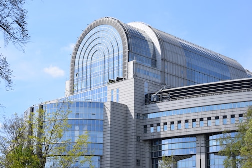 A modern architectural building with a curved glass dome structure on the roof. The facade consists predominantly of blue-tinted glass panels, complemented by gray concrete and metal elements. The surrounding area has some green trees and a clear blue sky.