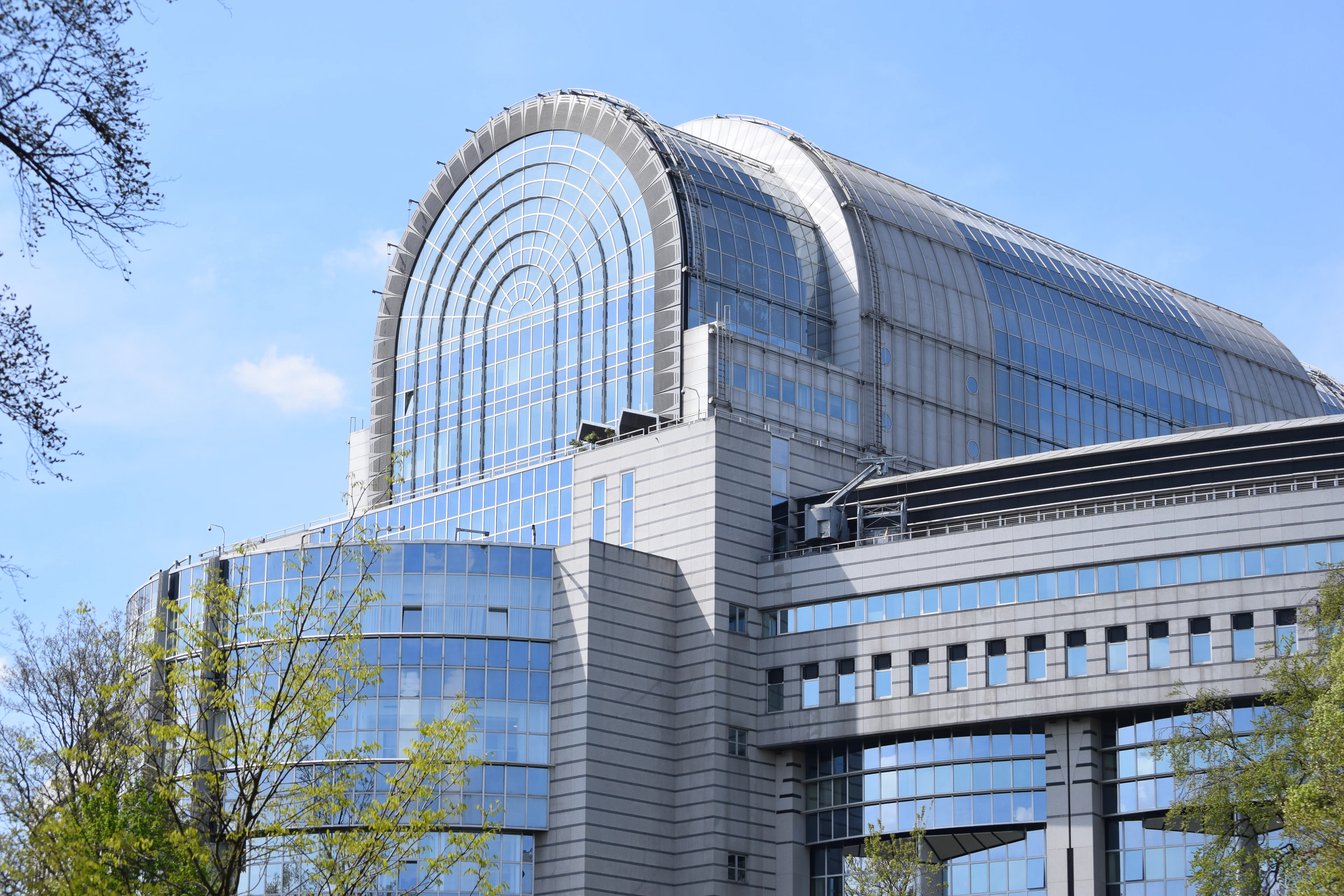 The curved glass facade of the European Parliament building in Brussels under a blue sky.